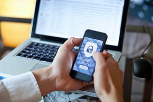 person holding phone with MFA security lock on screen and laptop in the background